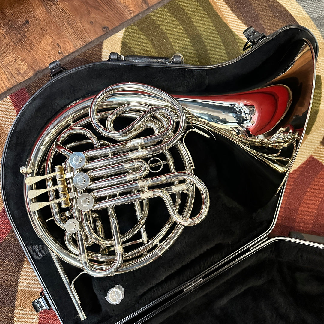 A silver French horn resting inside an open black instrument case on a patterned rug with a wooden floor visible at the side.