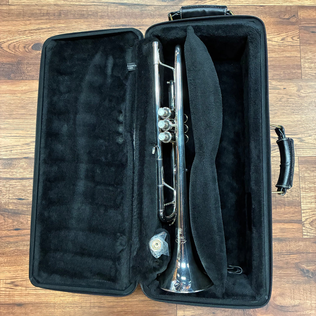 A silver trumpet resting inside an open black hard case with a black neck pillow, placed on a brown wooden floor.