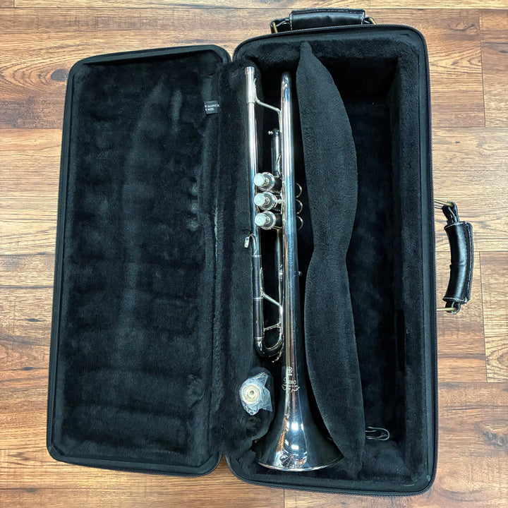A silver trumpet resting inside an open black hard case with a black neck pillow, placed on a brown wooden floor.